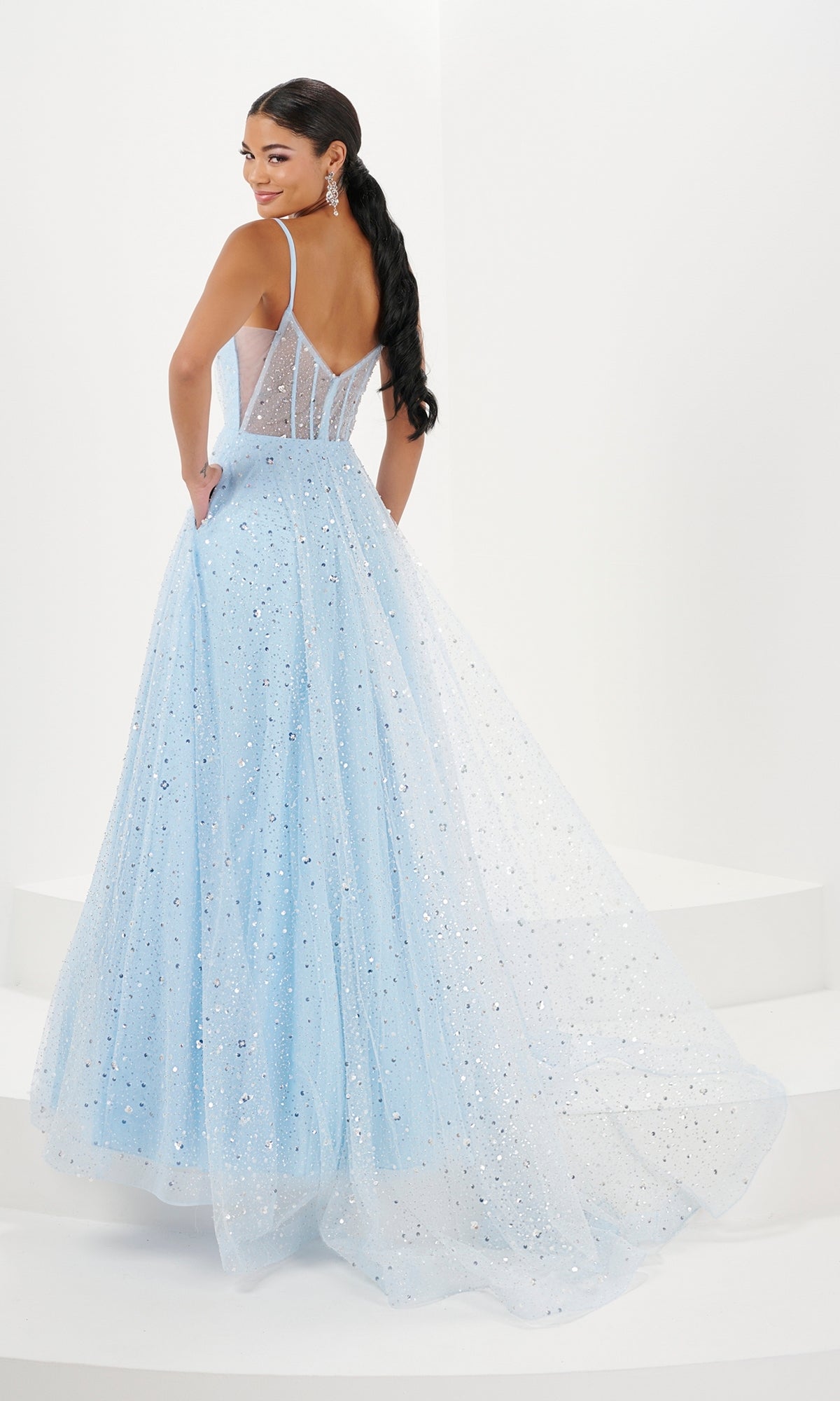 A woman in the Long Prom Dress 16066 by Tiffany—light blue, floor-length glitter tulle with thin straps, a sheer bodice, and v-neckline—stands on white steps, gazing over her shoulder.